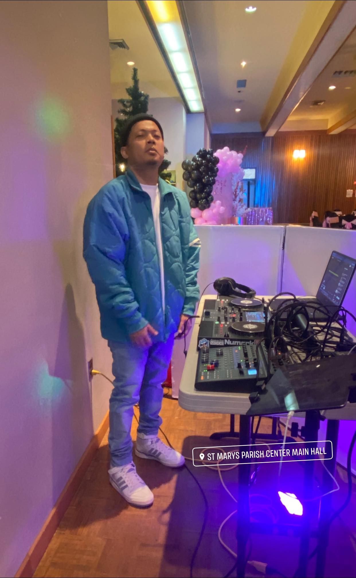 Person standing beside DJ equipment in a softly lit indoor venue with purple accent lights.
