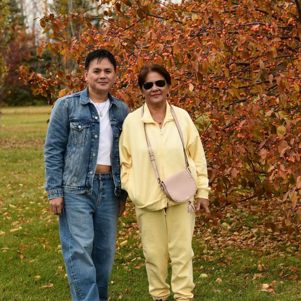 Two people standing in front of autumn foliage, one in denim and one in pale yellow clothing.