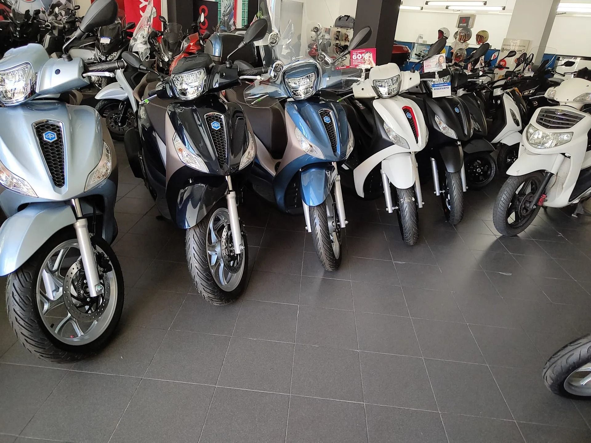 Fila di scooter in uno showroom; vari colori: blu, nero, bianco e argento.