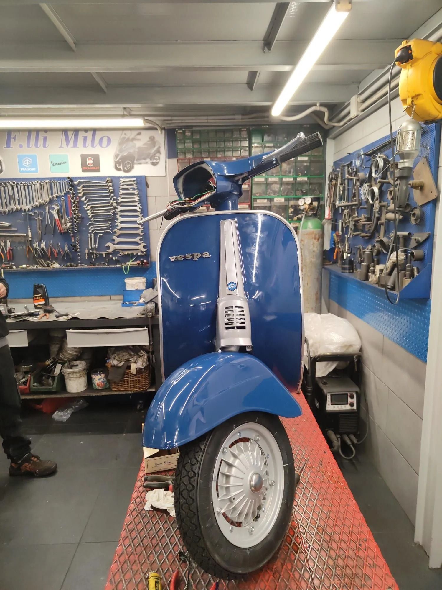 Una Vespa blu in un'officina, parcheggiata su un tavolo elevatore rosso. Gli attrezzi sono appesi alle pareti.