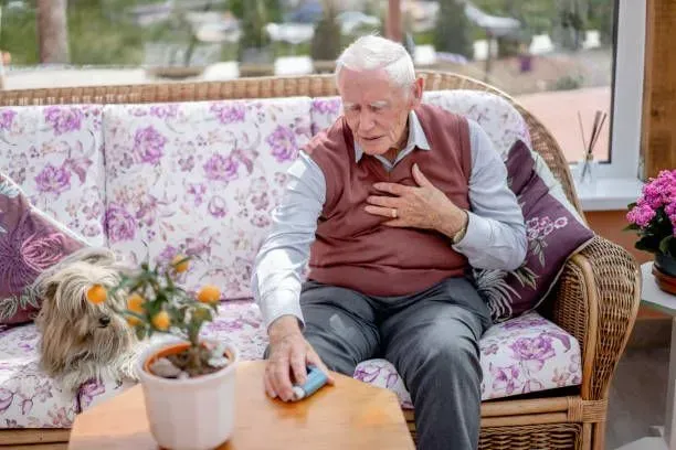An elderly man is sitting on a couch holding his chest.
