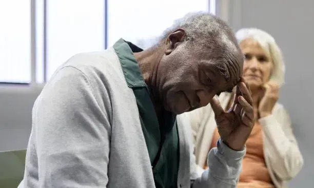 An elderly man is sitting in a chair with his head in his hand.