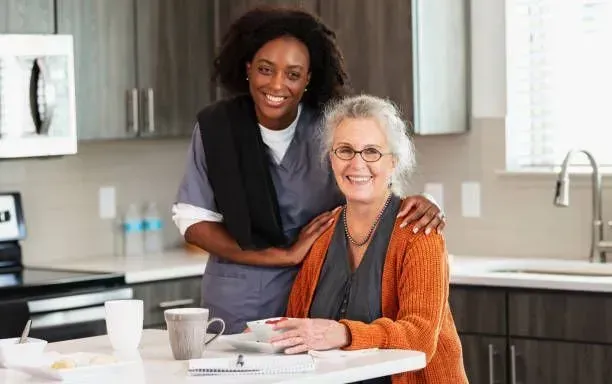 A nurse is standing next to an elderly woman sitting at a table in a kitchen.