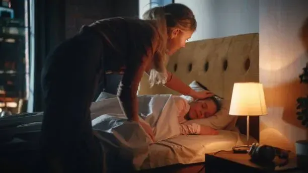 A woman is standing next to a man sleeping in a bed.
