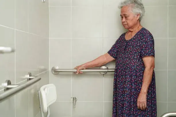 An elderly woman is standing in a bathroom holding onto a railing.