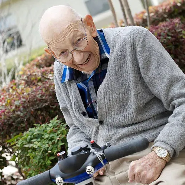 An elderly man wearing glasses is sitting on a scooter and smiling
