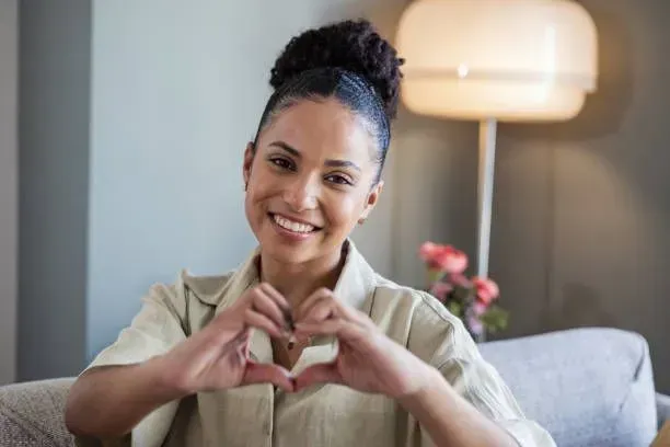 A woman is sitting on a couch making a heart shape with her hands.