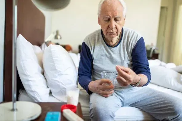 An elderly man is sitting on a bed taking a pill.