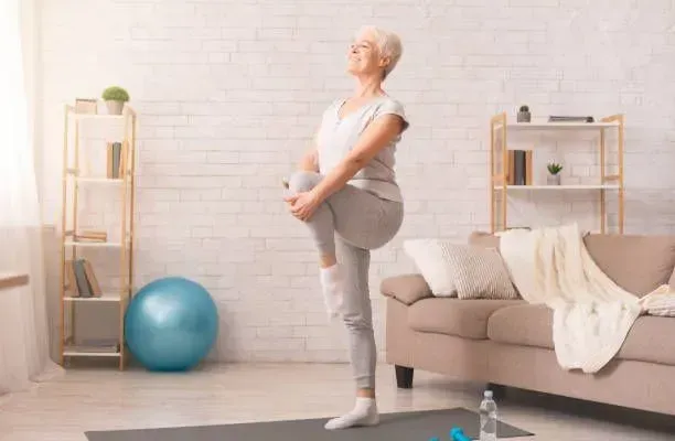 An elderly woman is stretching her legs on a yoga mat in a living room.
