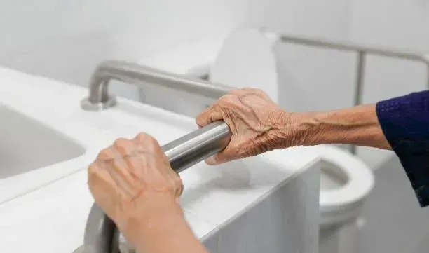 An elderly woman is holding onto a railing in a bathroom.