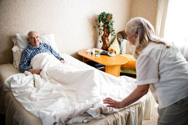 Elderly bedridden man in bed smiles at a caregiver, who straightens the covers in a bedroom.