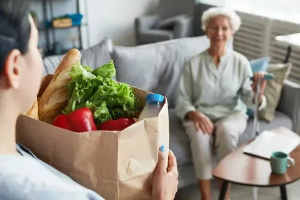 A woman is holding a bag of vegetables in front of an elderly woman sitting on a couch.