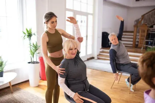 A woman is helping an older woman stretch her arms while sitting in a chair.