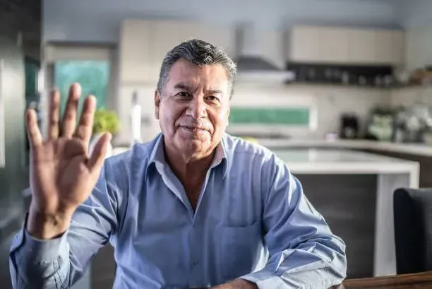 An older man is sitting at a table waving his hand.
