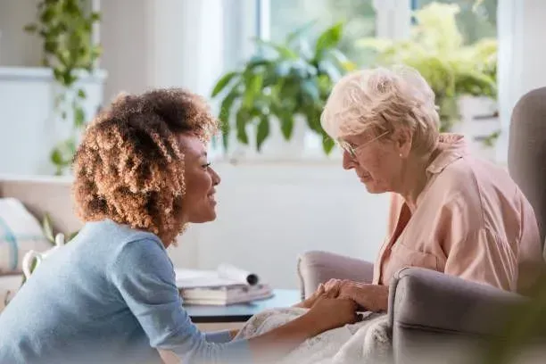 A woman is holding the hand of an older woman who is sitting in a chair.