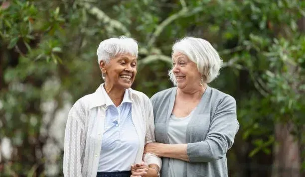 Two older women are standing next to each other in a park holding hands and smiling.