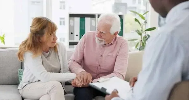 An elderly couple is sitting on a couch holding hands while talking to a doctor.