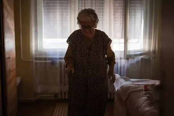 An elderly woman is standing in a dark room with a cane.