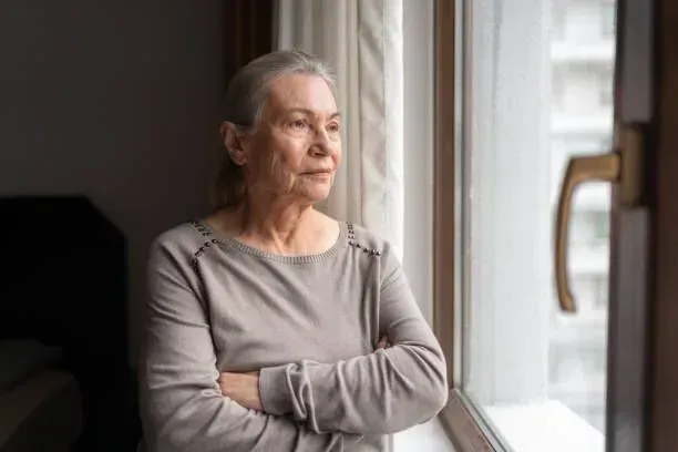 An elderly woman is looking out of a window with her arms crossed.