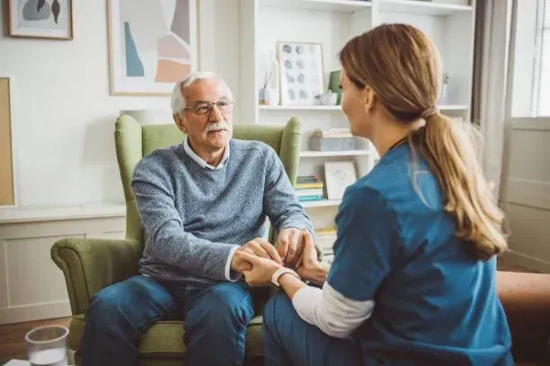A nurse is holding the hand of an elderly man while sitting in a chair.