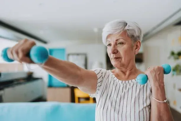 An elderly woman is lifting dumbbells in a living room.