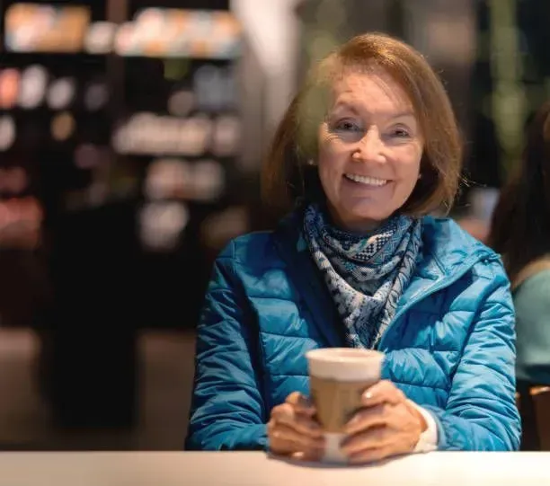 A woman in a blue jacket is sitting at a table holding a cup of coffee.