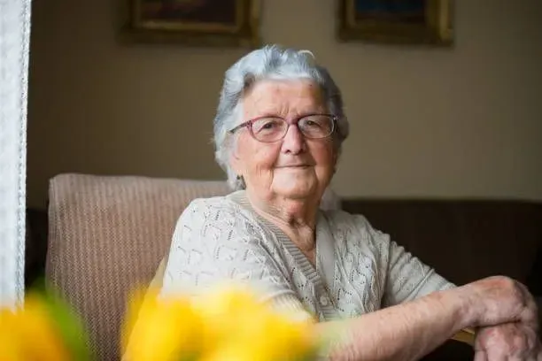 An elderly woman is sitting in a chair in front of a window.