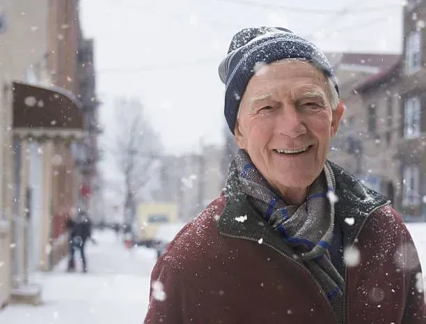 An elderly man wearing a hat and scarf is standing in the snow.