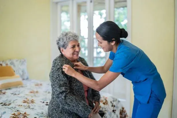 A nurse is helping an elderly woman dress in a bedroom.