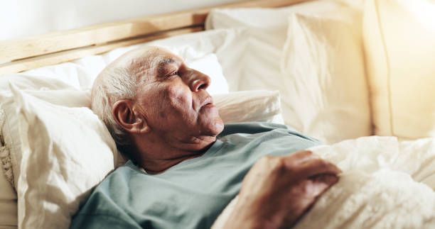 An elderly man sleeps peacefully in a bed, illuminated by sunlight.