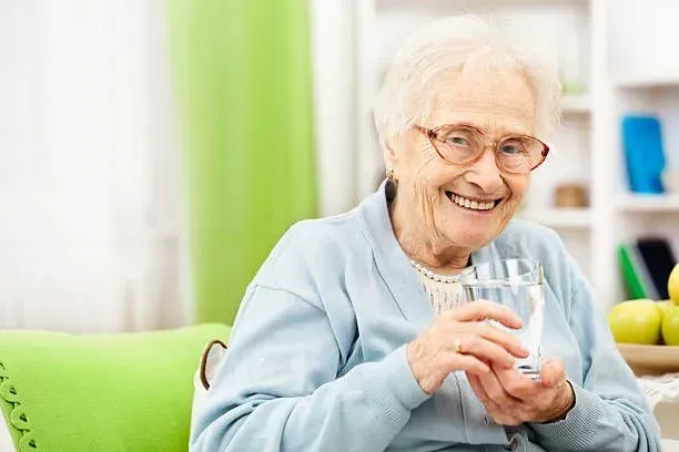 An elderly woman is sitting on a couch drinking a glass of water.