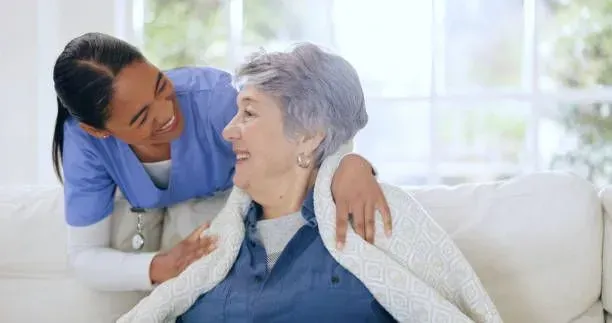 A nurse is putting a blanket on an elderly woman sitting on a couch.