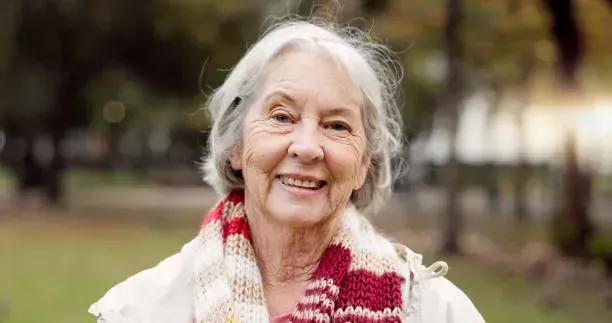 An elderly woman wearing a scarf is smiling in a park.