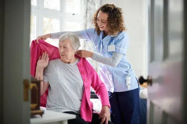 A nurse is helping an elderly woman dress in a bedroom.