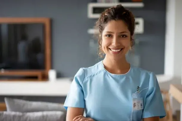 A nurse is smiling and standing in a living room with her arms crossed.