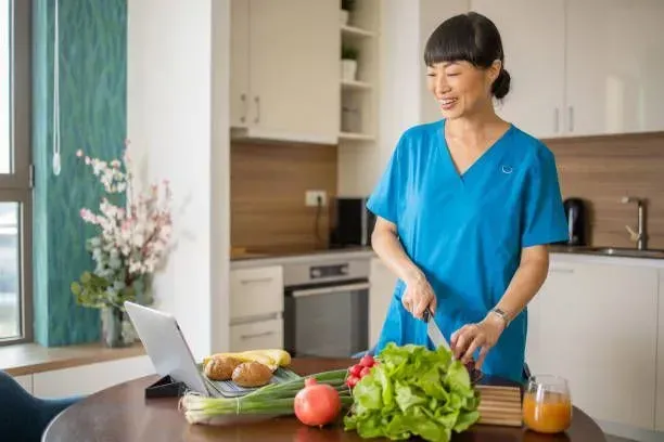 A woman is cutting vegetables in a kitchen while looking at a laptop.