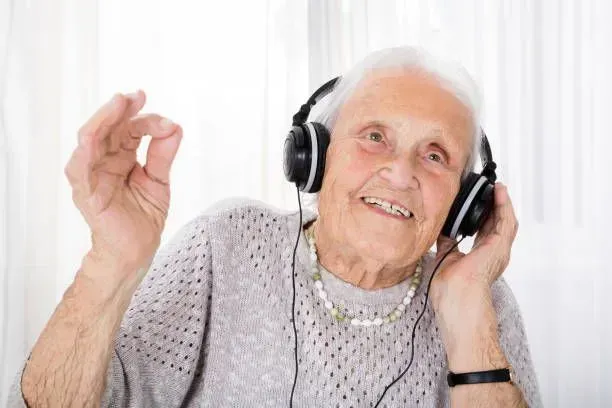 An elderly woman is wearing headphones and smiling.