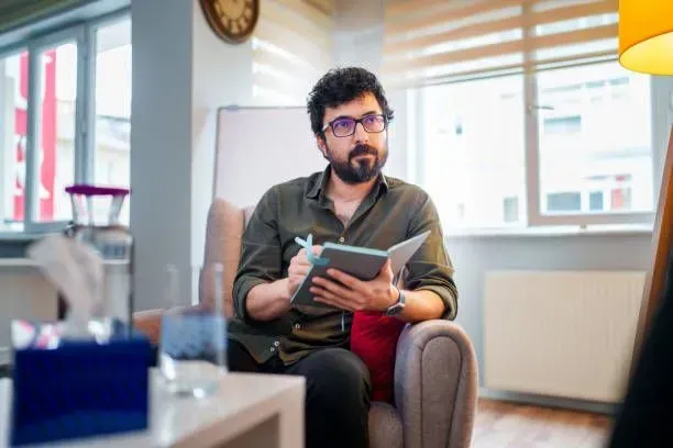 A man is sitting in a chair holding a notebook and a pen.