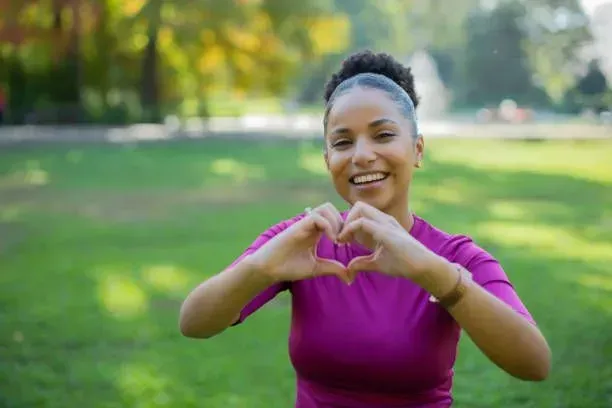A woman is making a heart shape with her hands in a park.