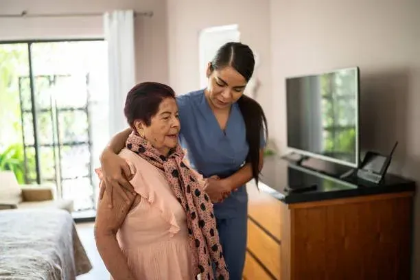 A nurse is helping an elderly woman walk in a bedroom.