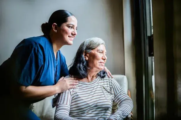 A nurse is standing next to an elderly woman looking out of a window.