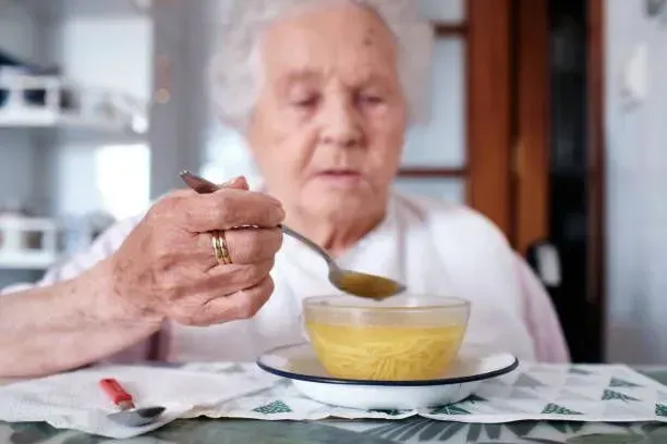 An elderly woman is sitting at a table eating from a bowl with a spoon.
