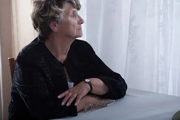 An elderly woman is sitting at a table looking out a window.