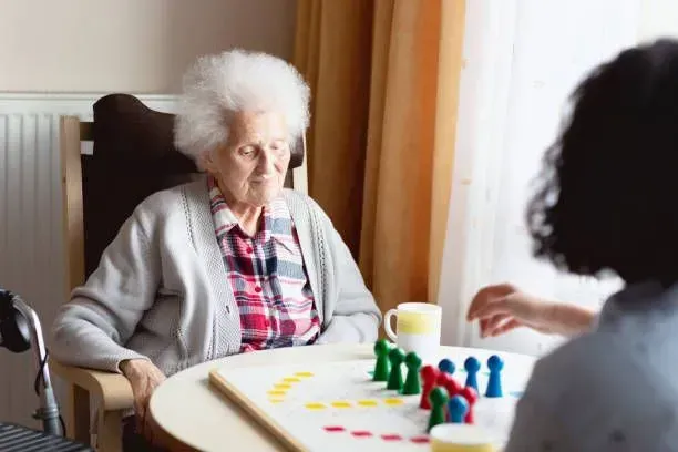 An elderly woman is sitting at a table playing a board game with a nurse.