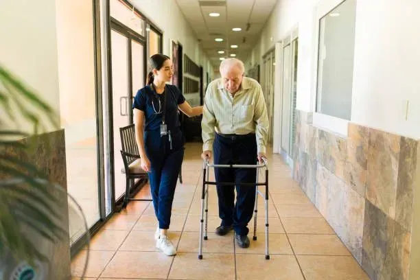 A nurse is helping an elderly man with a walker in a hospital hallway.