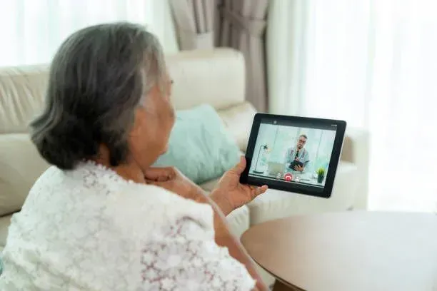 An elderly woman is sitting on a couch holding a tablet and talking to a doctor.