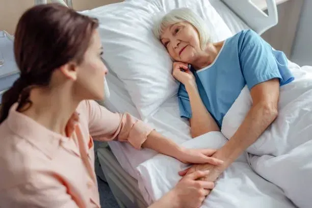 A woman is holding the hand of an elderly woman in a hospital bed.