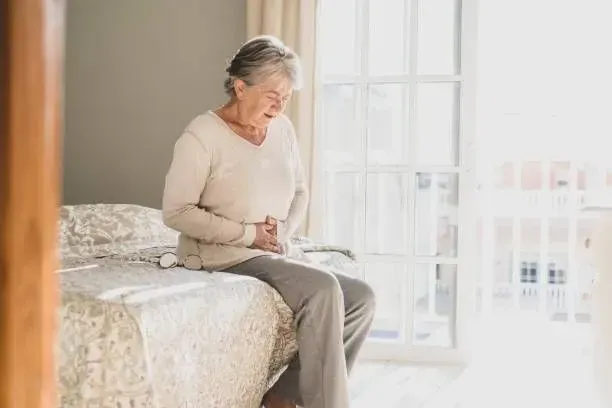 An elderly woman is sitting on a bed holding her stomach.
