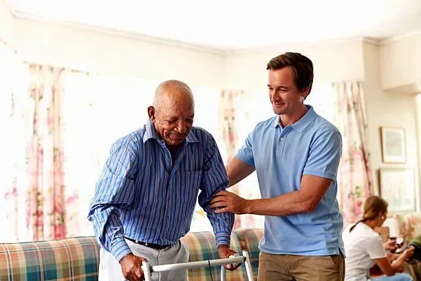 A man is helping an older man with a walker in a living room.