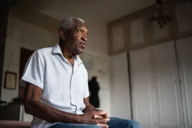 An elderly man is sitting on a bed in a room with his hands folded.
